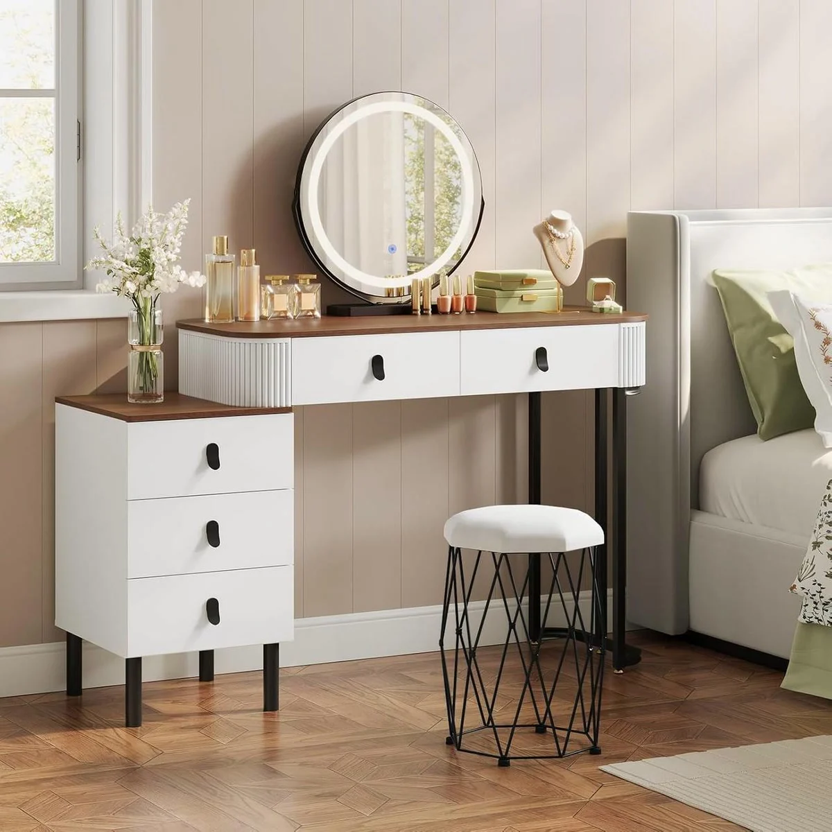 Tocador de maquillaje de madera y blanco en un dormitorio, con espejo redondo con luz, cómoda y taburete de terciopelo